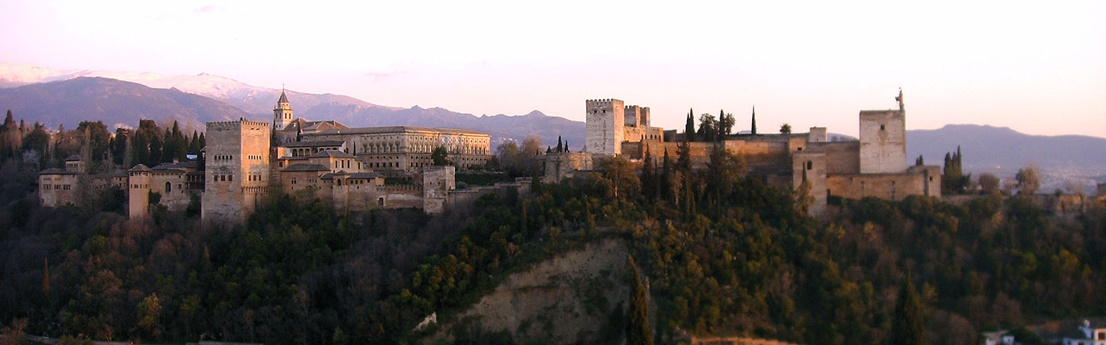 The Alhambra fortress, viewed from a distance at sunset, with mountains in the background.